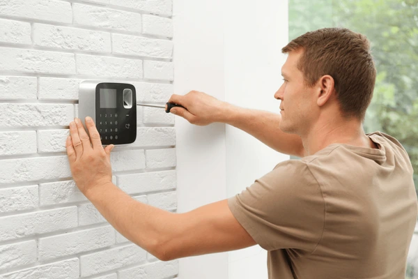 Male technician installing a security alarm system on a white brick wall, emphasizing professional access control solutions for enhanced home and business security.